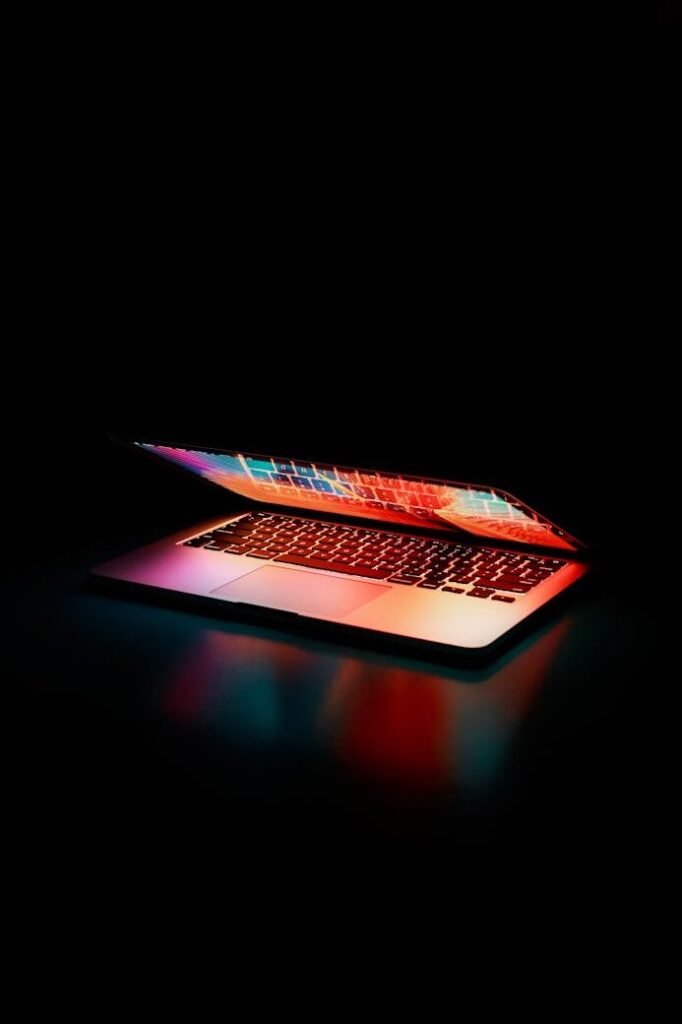 Laptop partially opened, emitting vibrant multicolored lights in dark setting.